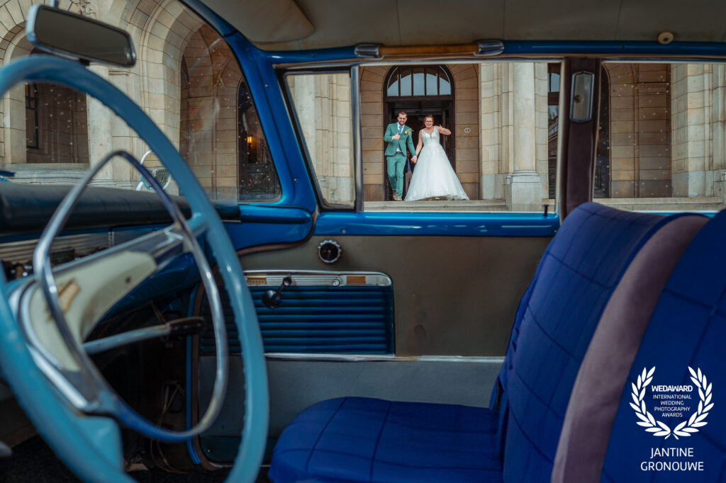 Bruidspaar komt hand in hand de trap af, gefotografeerd door de voorruit van een blauwe oldtimer. Winnaar van een WedAward.