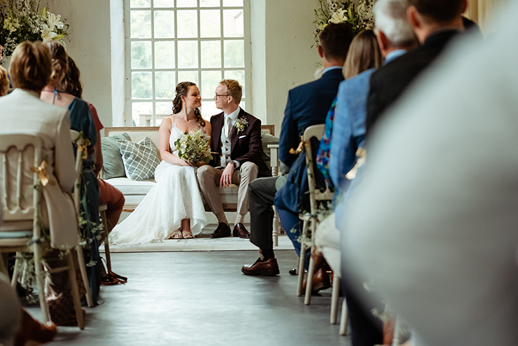 Bruidspaar zit tussen de gasten in de zaal met hoge ramen tijdens de ceremonie op Landgoed Klarenbeek