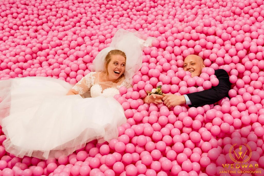 Bruid in witte jurk en bruidegom liggen lachend in een roze ballenbak. Wedwar Award.