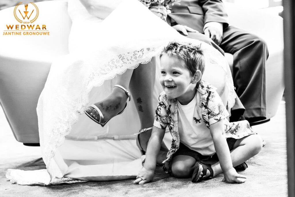Zwart-witfoto: een kind kruipt lachend onder de trouwjurk van de bruid vandaan tijdens de ceremonie. Wedwar Award.