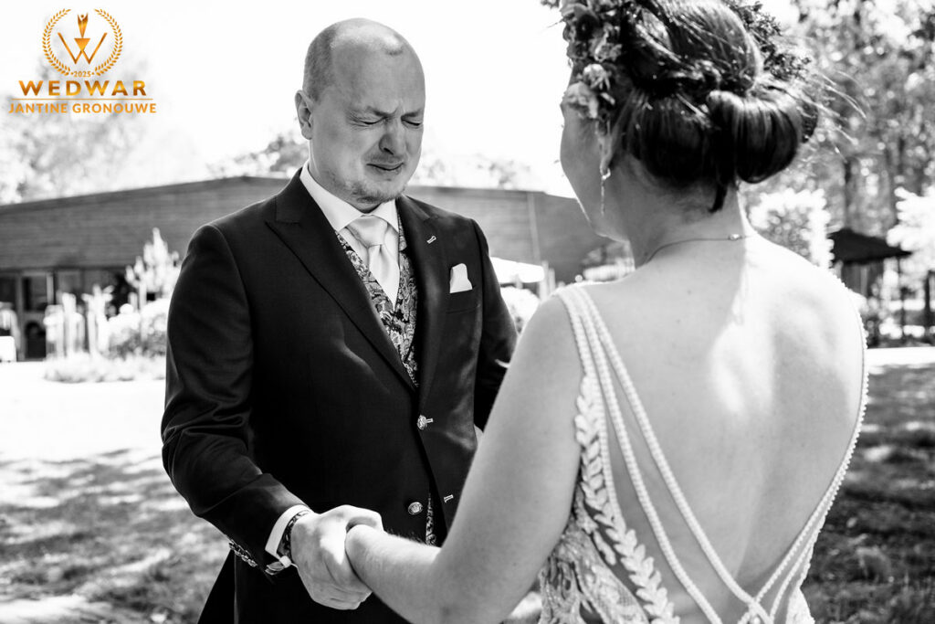 Zwart-witfoto van de first look: de bruidegom veegt een traan weg terwijl de bruid zijn hand vasthoudt. Wedwar Award.