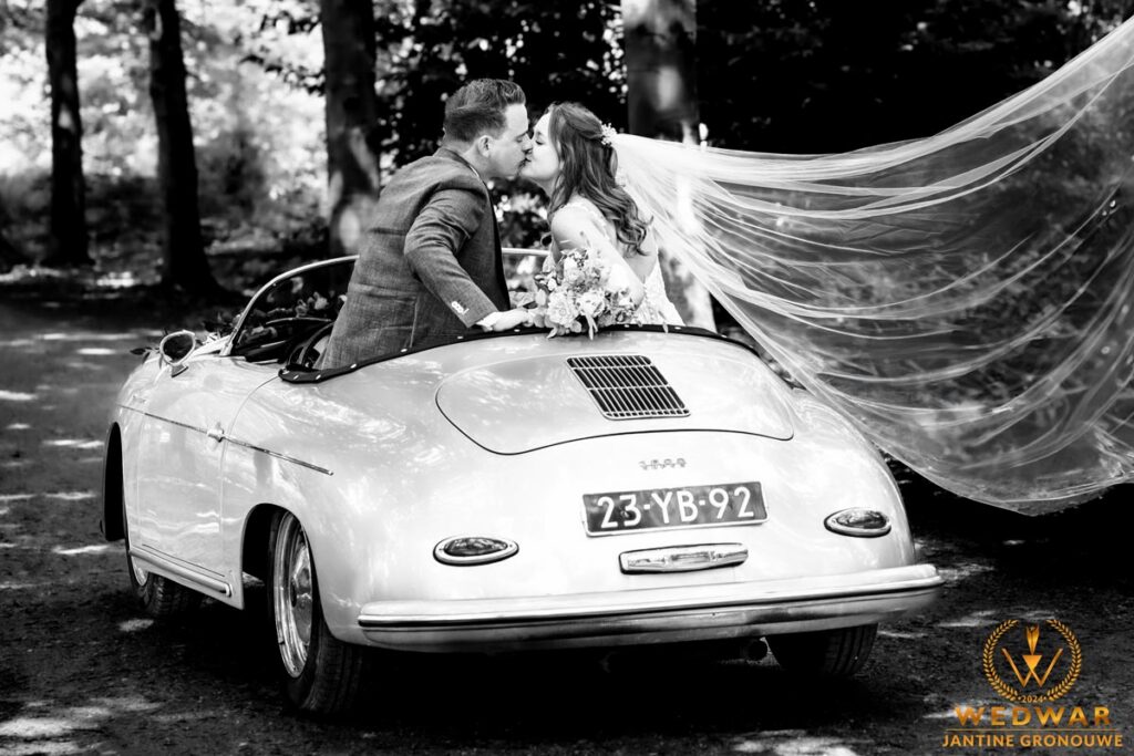 Zwart-witfoto: bruidspaar kust elkaar in een oude Porsche cabrio terwijl de sluier van de bruid opwaait. Wedwar Award.